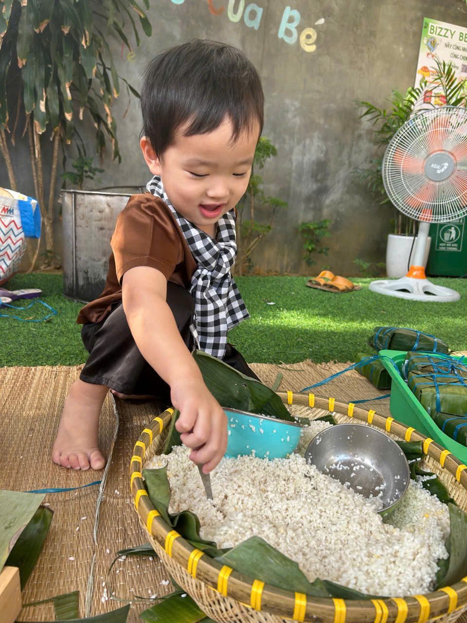 A young child with black hair, wearing a brown shirt, dark pants, and a black-and-white checkered scarf, crouches on a woven mat while playing with rice in a large basket lined with green leaves. The child appears happy and engaged, with a backdrop o