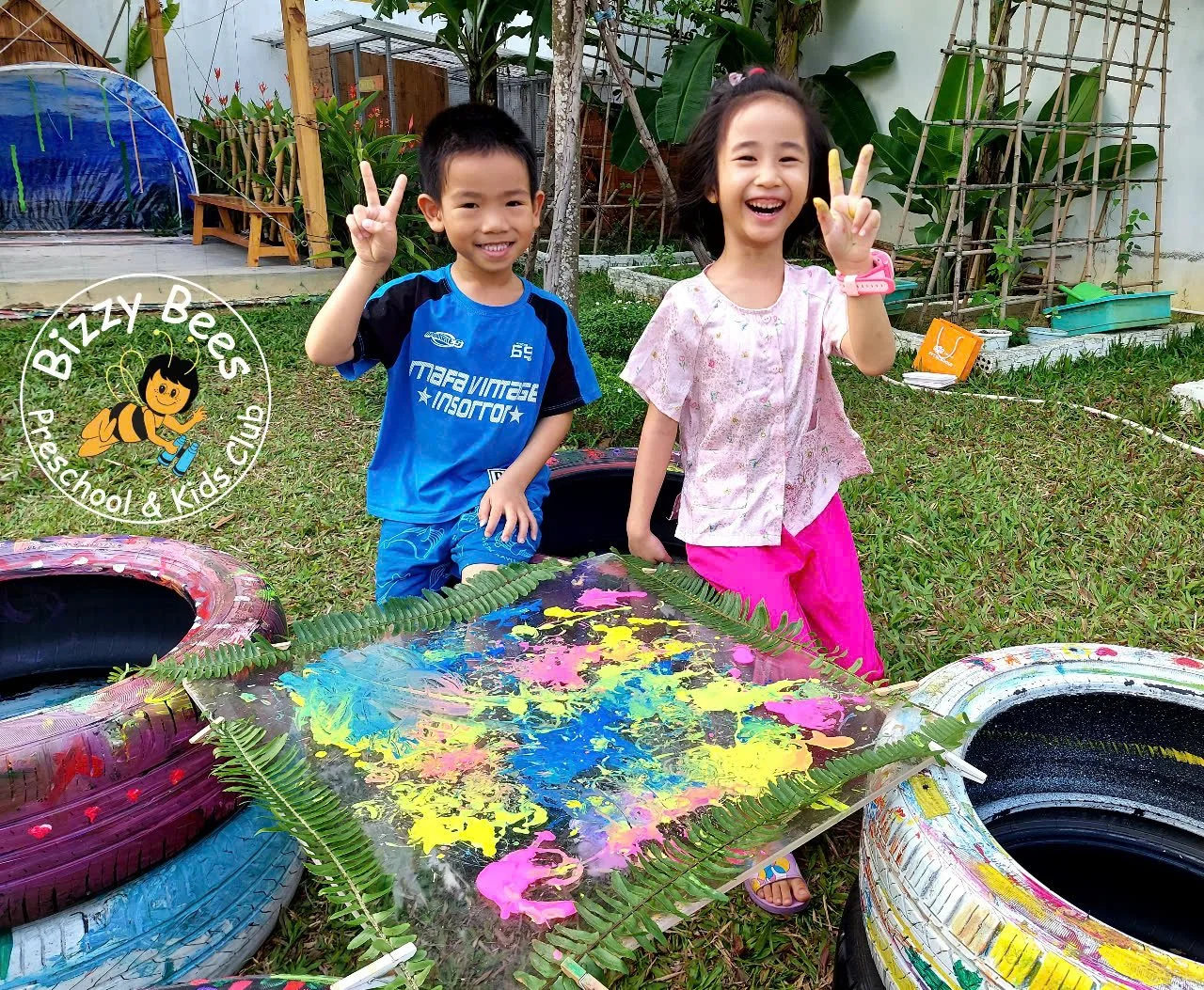 Two children, a boy and a girl, smiling and making peace signs with their fingers, standing beside a colorful painting on a table outdoors in a garden with plants and tires arranged creatively.
