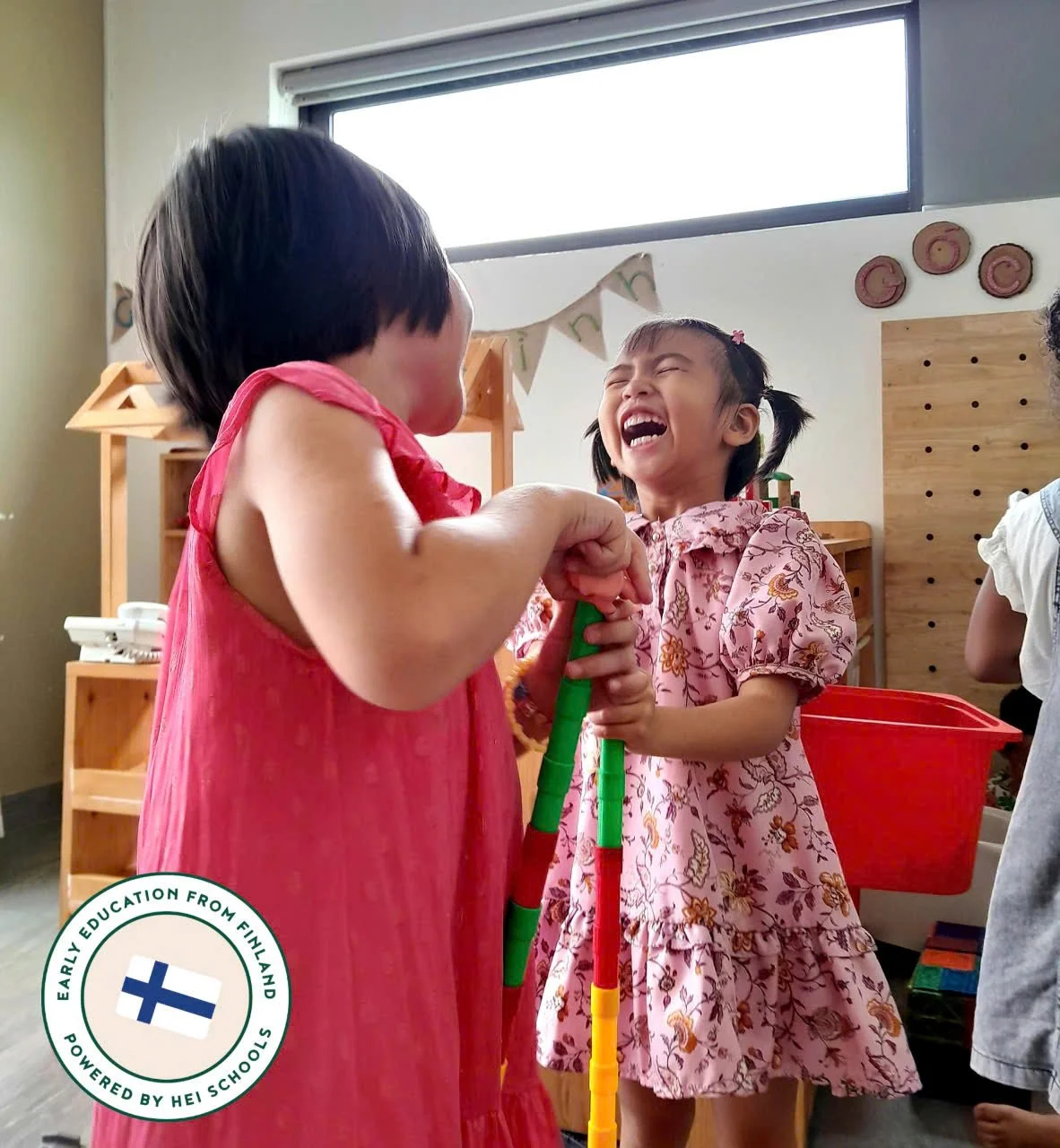 Two young girls laughing and playing with a colorful toy stick in a classroom, with wooden shelves and wall decorations in the background.