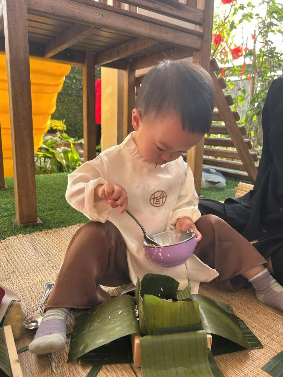A young child sitting cross-legged on the floor, eating from a purple bowl with a spoon, surrounded by green banana leaves and a woven mat, with a wooden play structure and plants in the background.