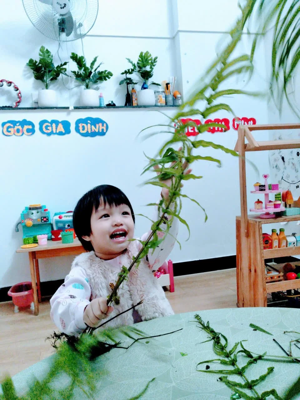 A young child with short black hair, smiling and holding a green leafy plant, sitting at a table in a classroom or playroom setting.