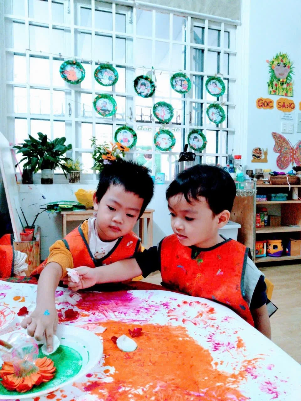 Two young boys with paint-smeared hands decorating a cake at a table in a classroom. The background includes colorful holiday decorations, plants, and shelves with toys and supplies.