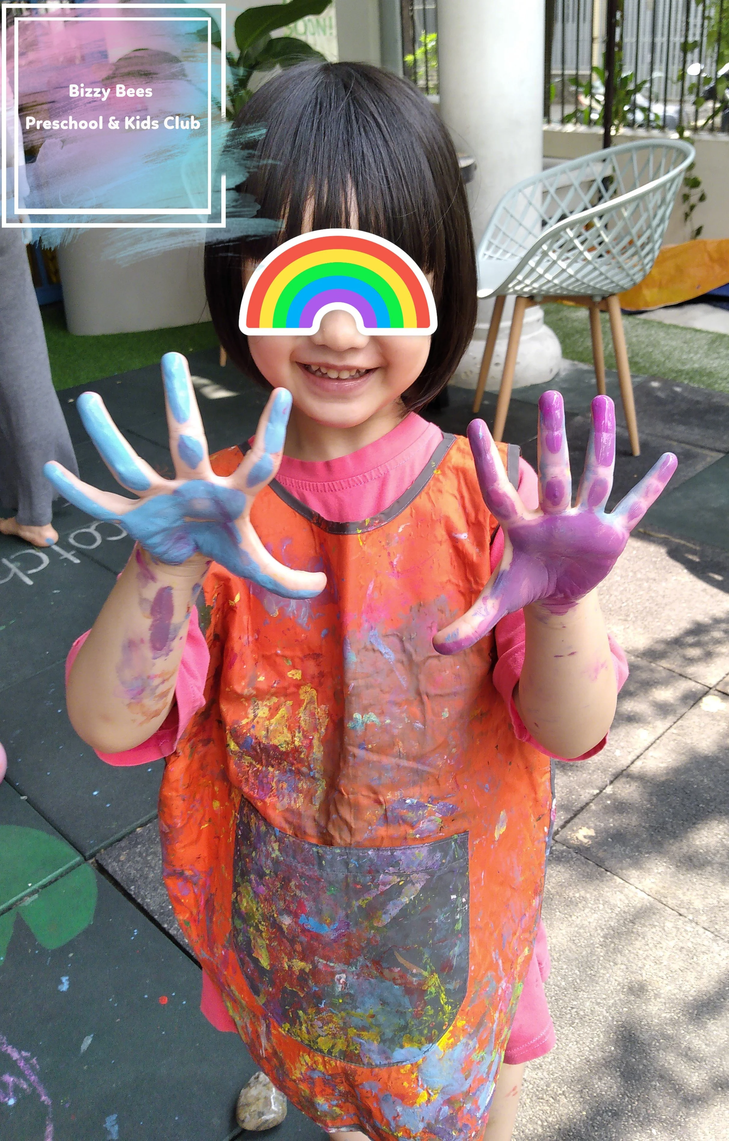 A young girl smiling with painted hands, wearing an orange apron, with a rainbow sticker over her eyes, at Bizzy Bees Preschool & Kids Club in Da Nang