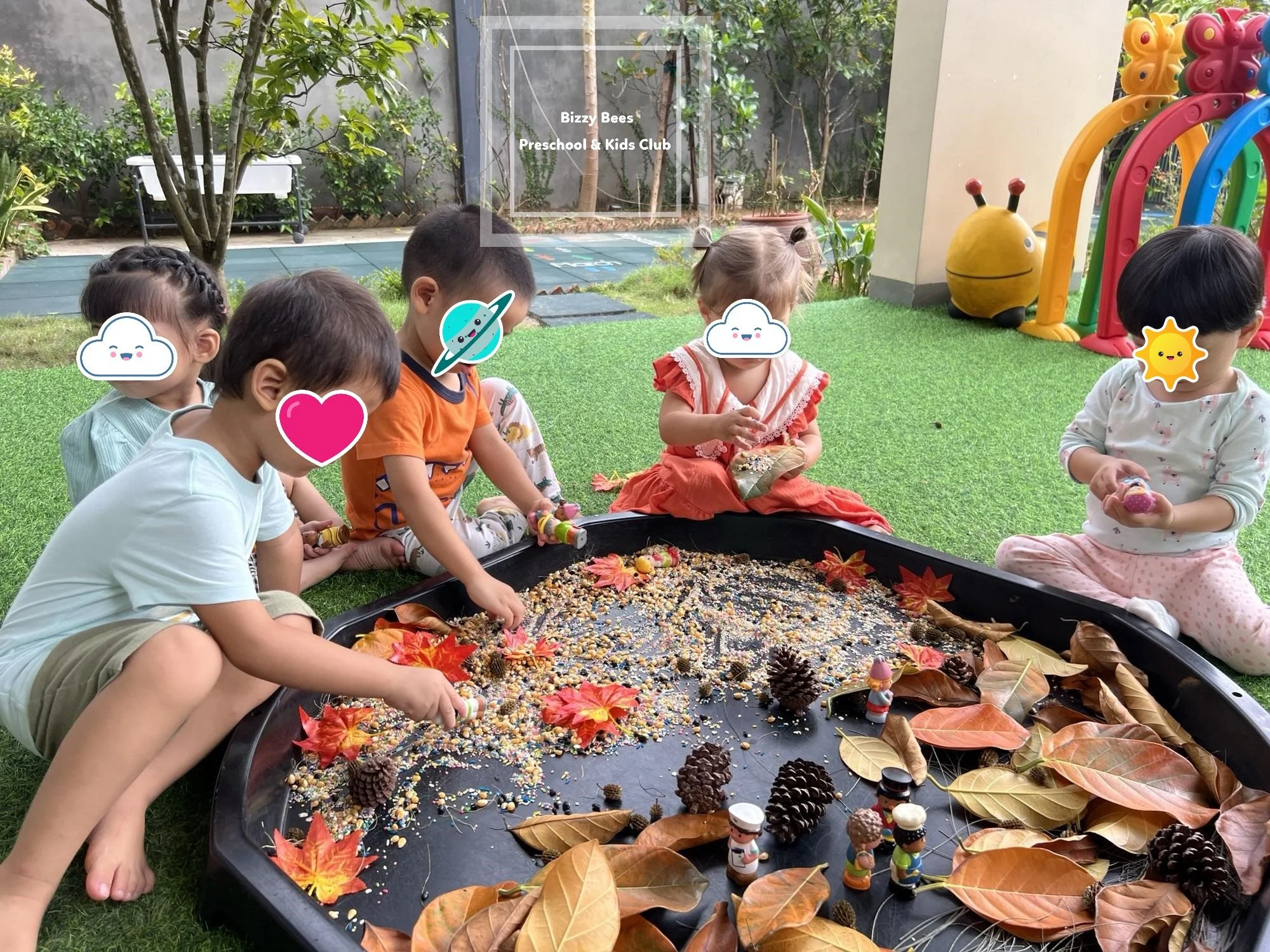 Children playing with colorful pretend food on an outdoor playground with autumn leaves and pinecones, supervised by a teacher at Bizzy Bees Preschool & Kids Club.