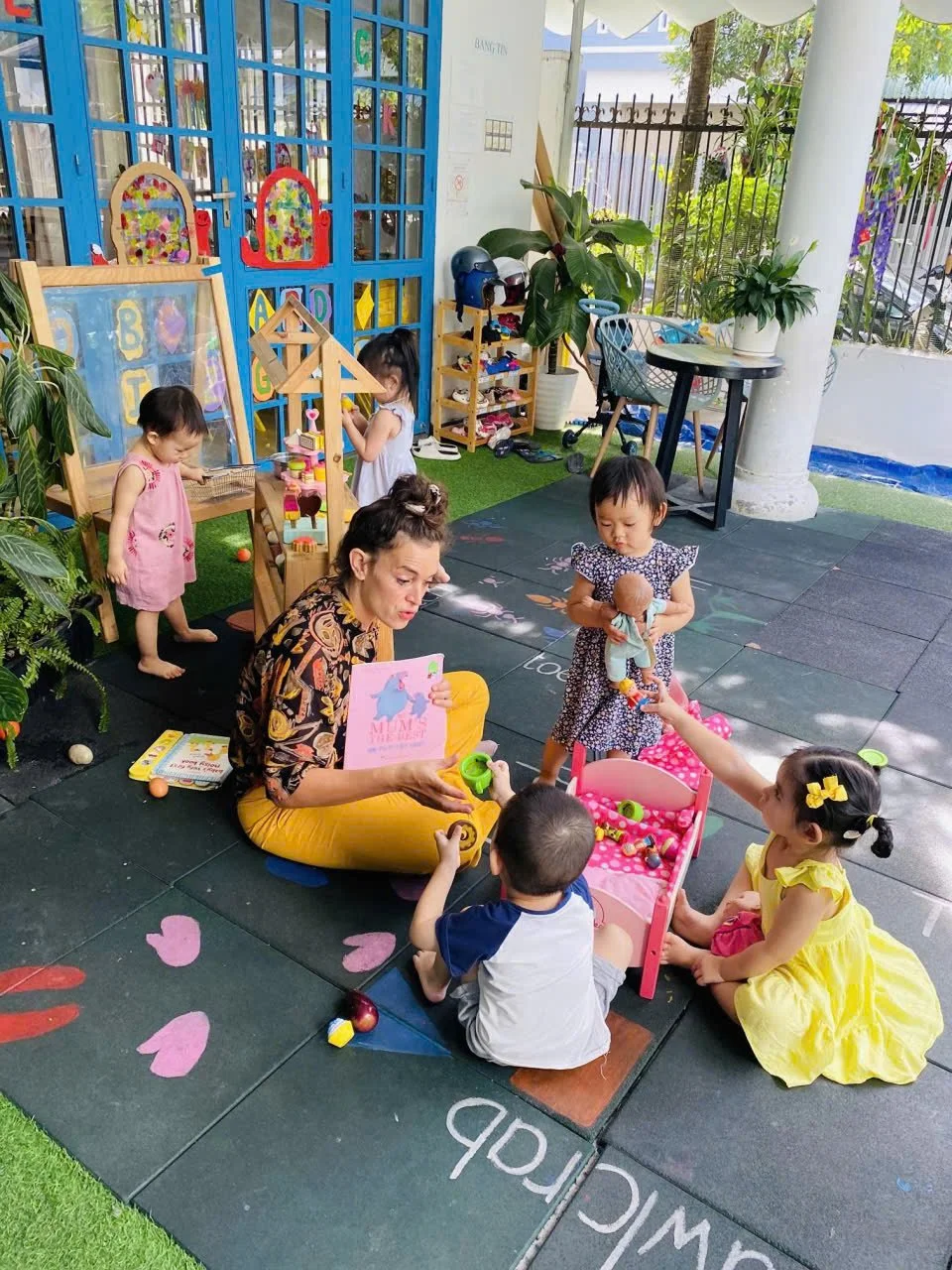 A woman and four young children playing on a black rubber mat outside a colorful building with a blue door. Children are engaged in various activities including playing with toys, dolls, and small objects. The area has green plants, and there's outdo