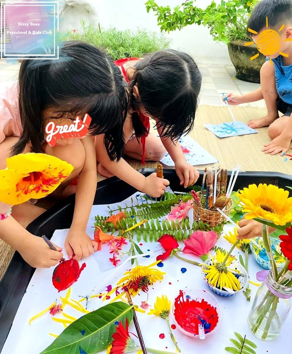 Children painting and crafting with flowers and leaves outdoors, with art supplies on a table, at Bizzy Bees Preschool & Kids Club.