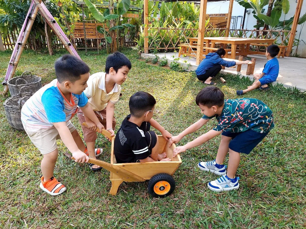 Group of children playing outdoors with a wooden wagon, some pulling and one sitting inside while others push. Two children sit on a picnic table in the background, with greenery and a wooden structure behind them.