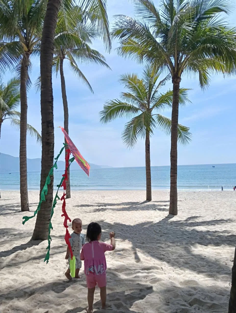 Children playing under palm trees on a sandy beach with blue ocean and distant mountains in the background.