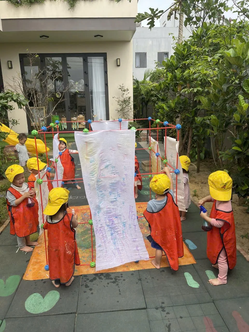 Four children are painting on a large white board in an outdoor playground area. One child is standing and painting, while the other three are sitting or kneeling, working on the board with various colors of paint.