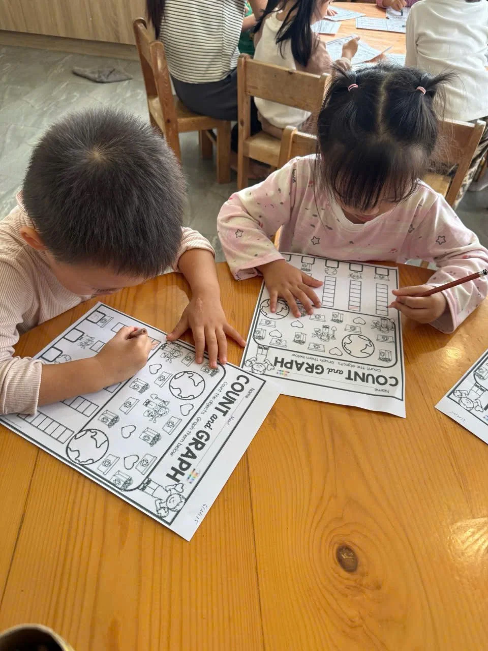 Two children sitting at a wooden table, coloring and working on a counting and geography activity sheet, with other children and adults in the background.