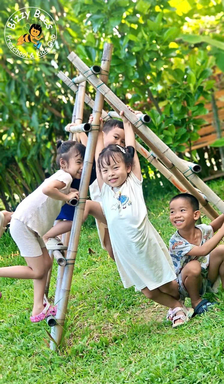 Four young children playing on a bamboo jungle gym outdoors, surrounded by green foliage and grass, smiling and having fun.