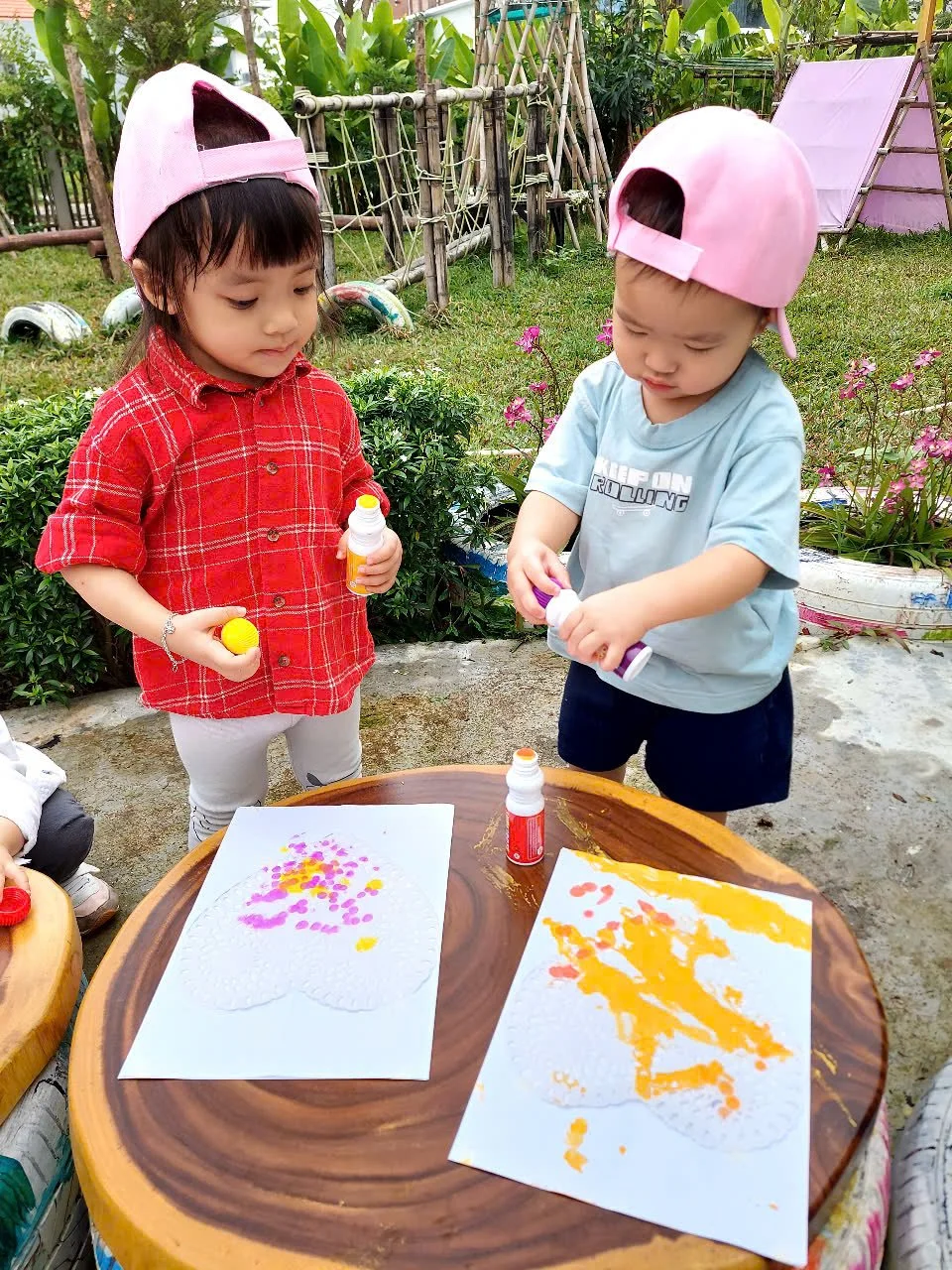 Two young children, a girl in a red plaid shirt and a boy in a light blue shirt, wearing pink caps, are engaging in arts and crafts outdoors. They are standing at a round wooden table with white paper, colorful paint, and bottles of glue. The girl ho