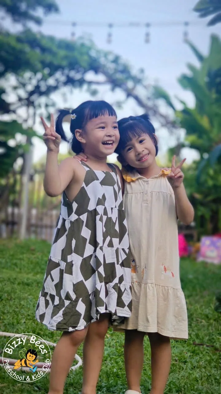Two young girls smiling and making peace signs with their hands, standing outdoors on grass with trees and string lights overhead in the background.