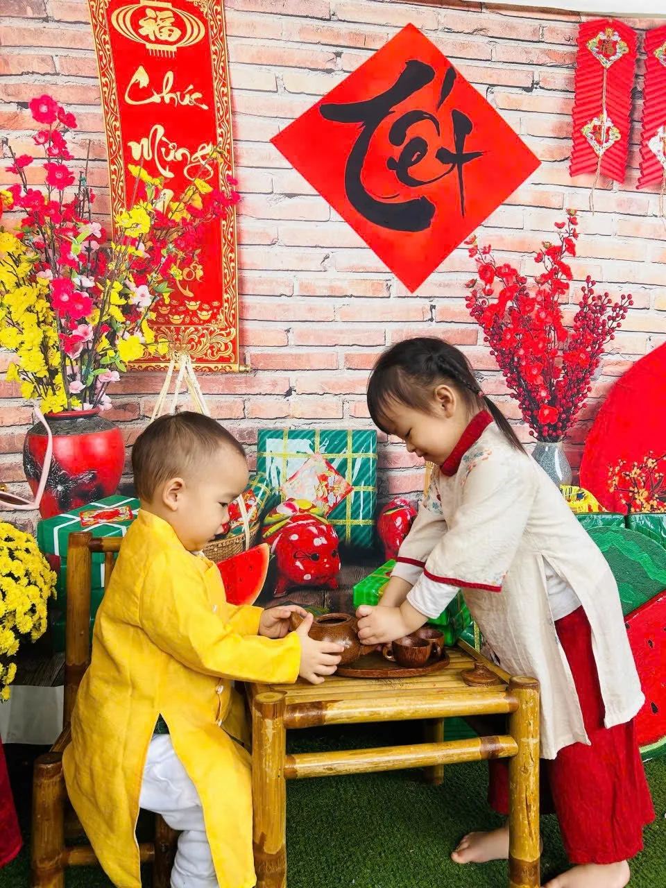 Two children, a young boy in a yellow traditional outfit and a girl in a cream and red outfit, are engaging in a tea ceremony with small clay cups on a wooden table. The background features red and yellow floral arrangements, Chinese calligraphy, and