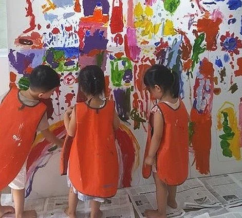 Three children painting on a large white wall with colorful abstract artwork at Bizzy Bees Kindergarten in Da Nang
