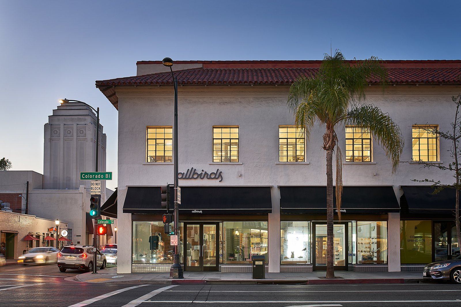 Front view of an Allbirds store on a corner with a street sign for Colorado Blvd, featuring a white building with a red-tiled roof and large windows, palm tree in front, and cars on the street.
