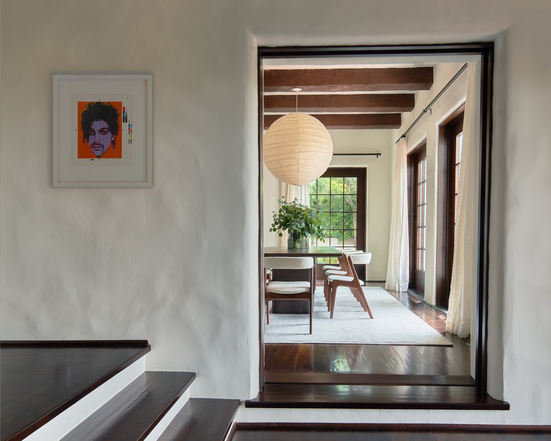 Interior of a modern dining room with a wooden table, chairs, and a large paper lantern hanging from the ceiling. The room has natural light coming through large windows with white curtains. There is a small piece of artwork on the adjoining wall.