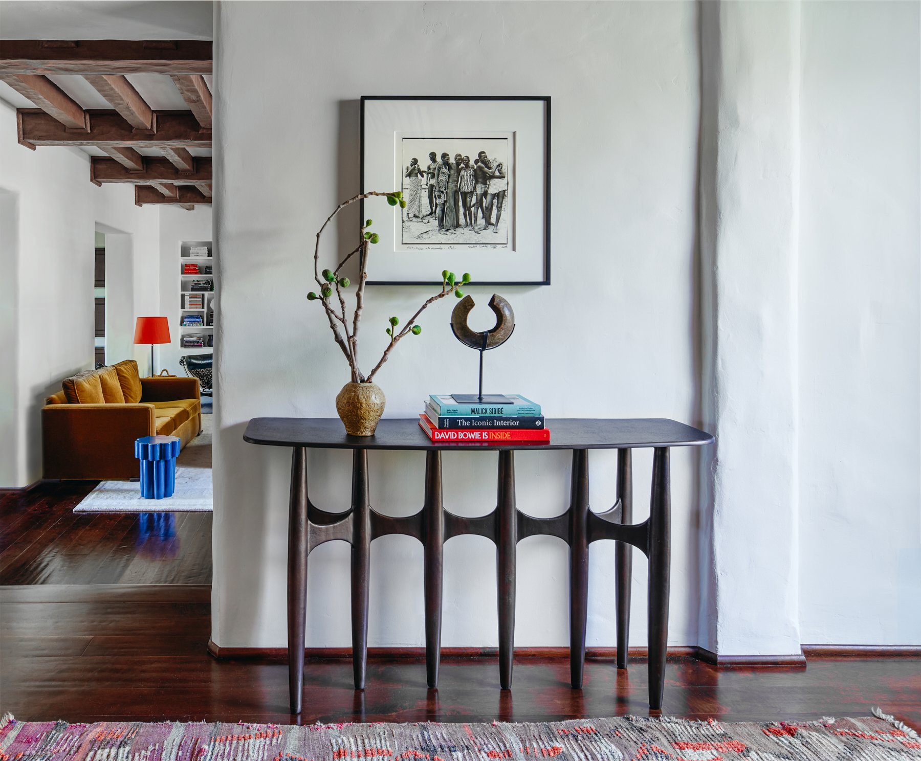 Stylish interior with a dark wooden console table adorned with books and decorative items, a framed artwork above, and a glimpse of a living room with a mustard sofa and colorful decor.