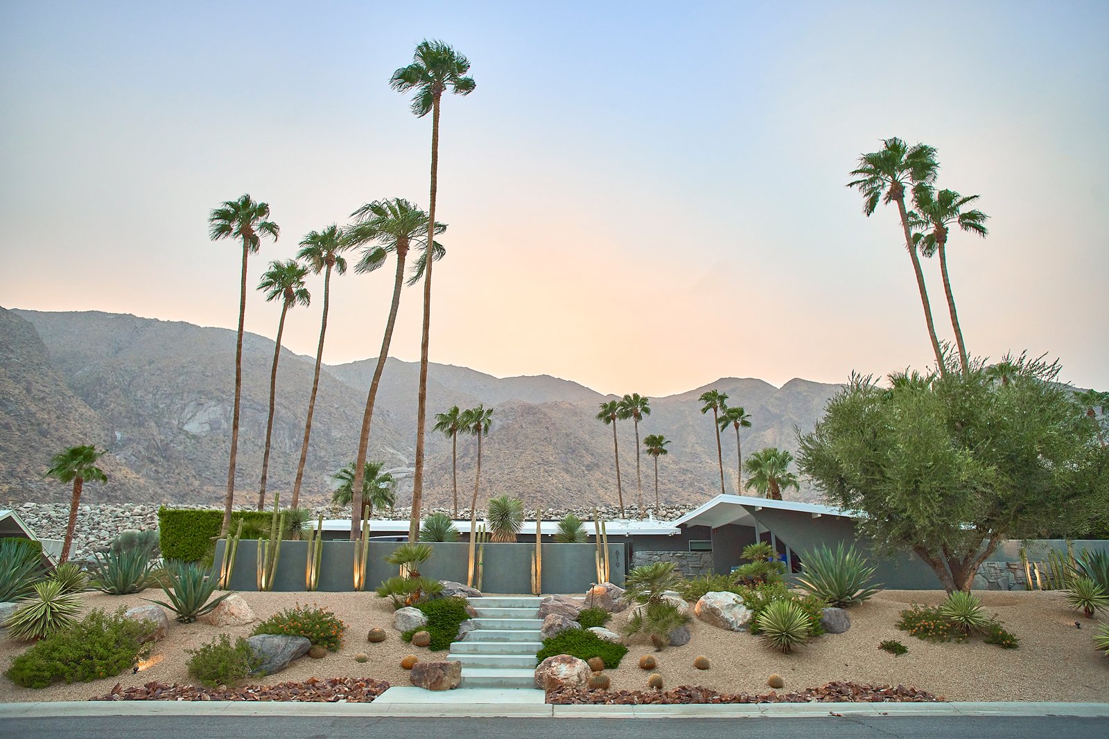 Mid-century modern house with desert landscaping and palm trees against mountain backdrop under a clear sky.