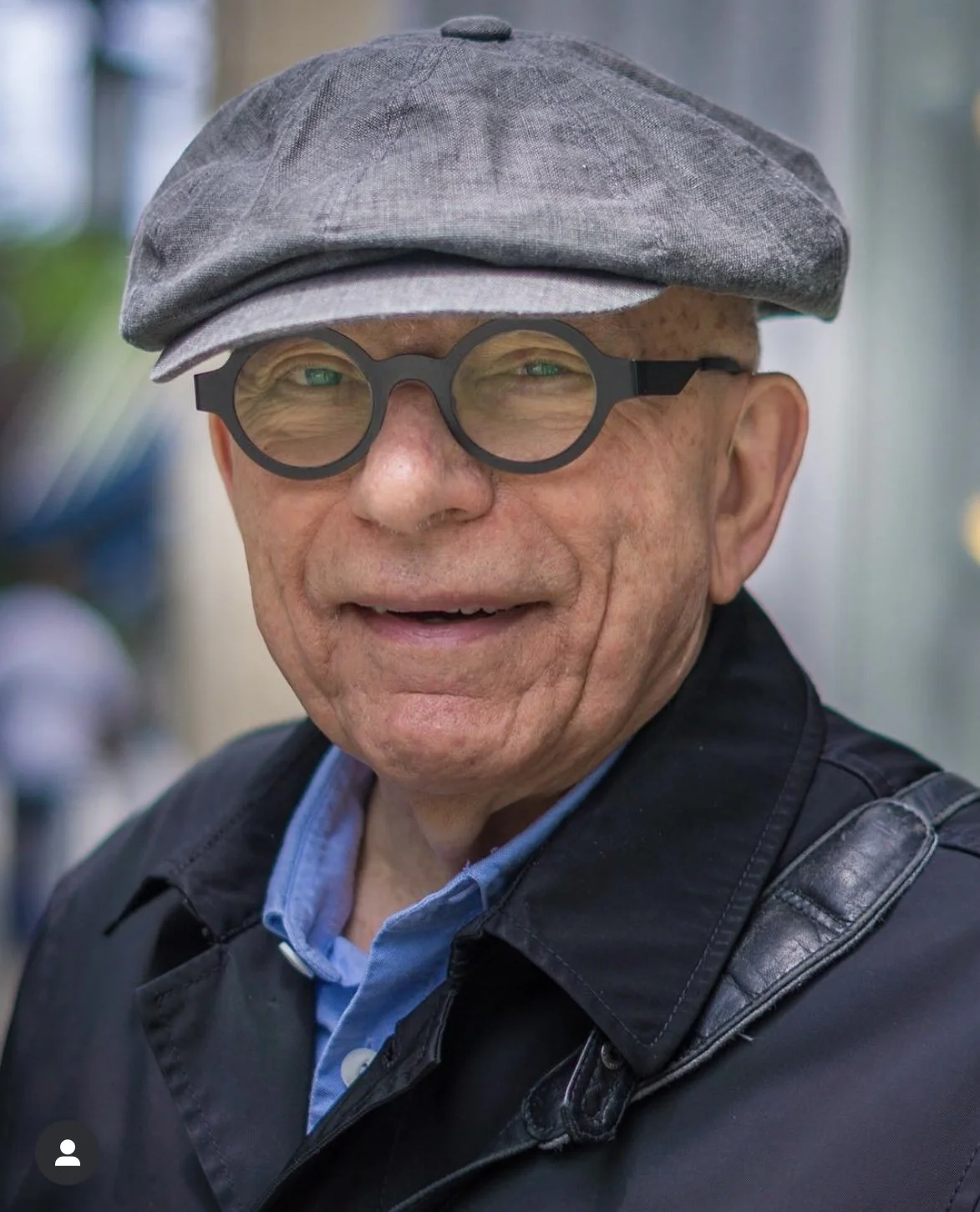 Elderly man wearing a flat cap and glasses, smiling, with an outdoor background.