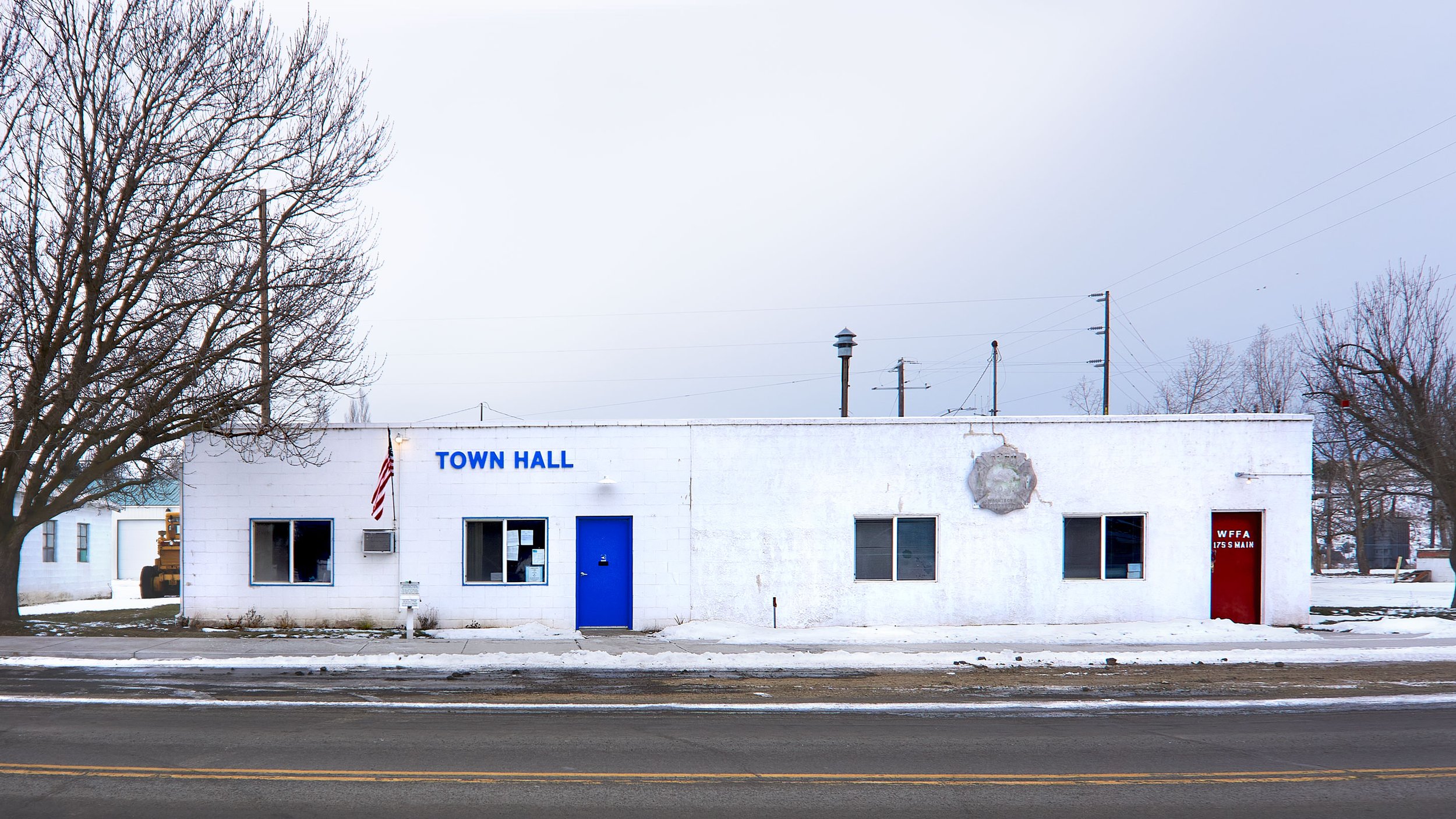 Small town hall building with an American flag, blue door, and snowy surroundings.