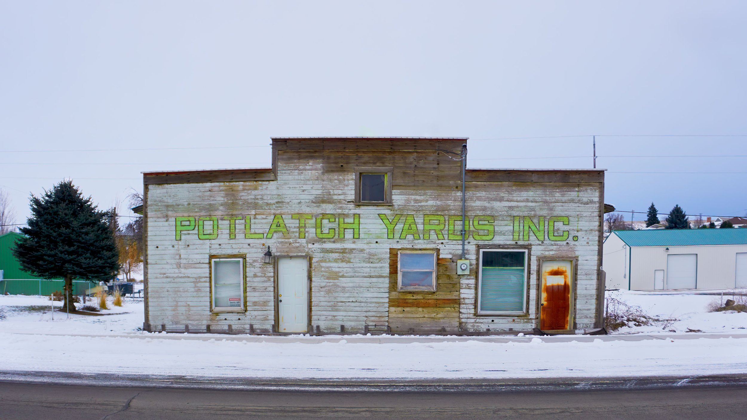 Old wooden building labeled "POTLATCH YARDS INC." with snowy ground and overcast sky.
