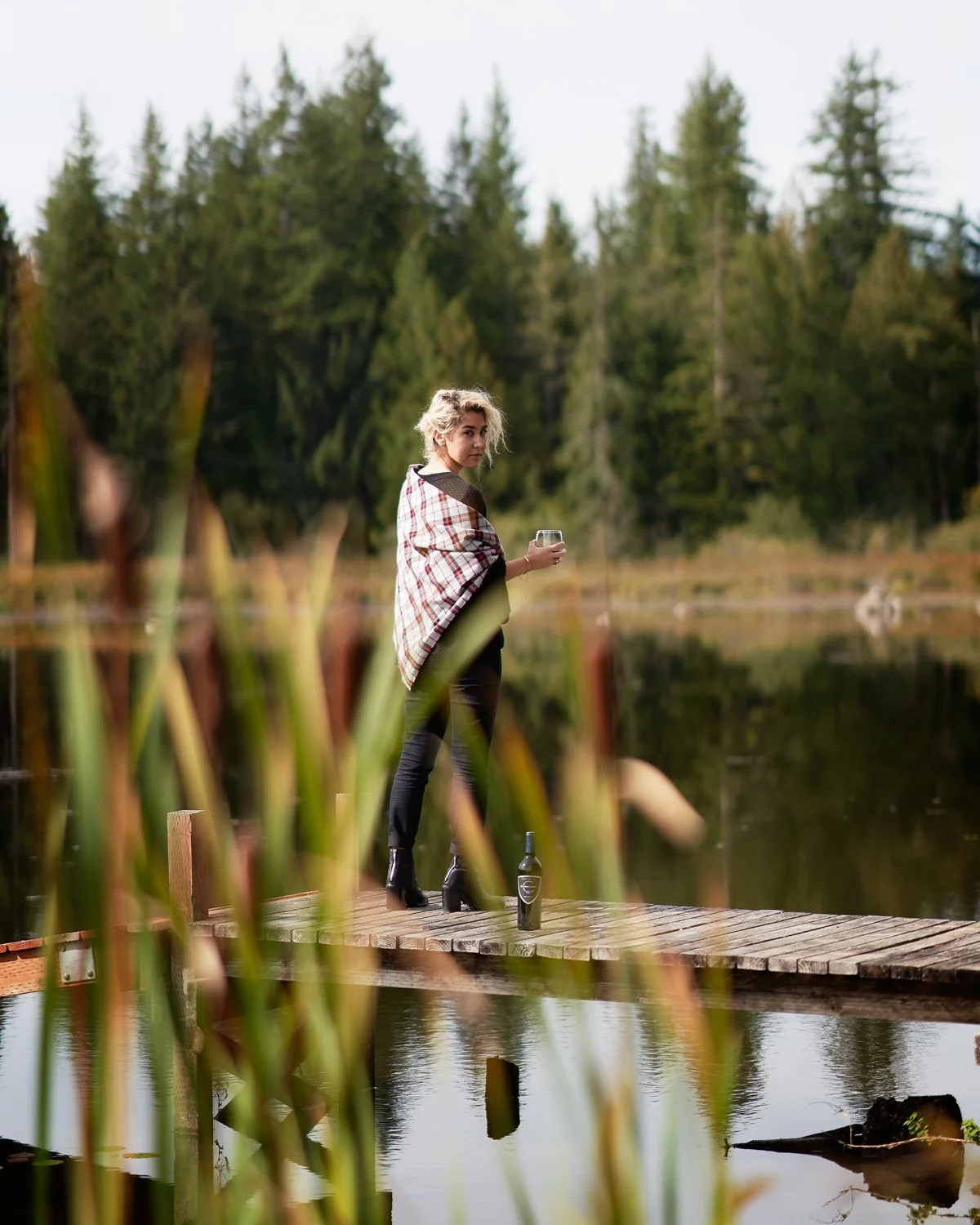 Person standing on a wooden dock by a lake, wrapped in a plaid blanket, holding a drink, with a wine bottle on the dock, surrounded by trees.