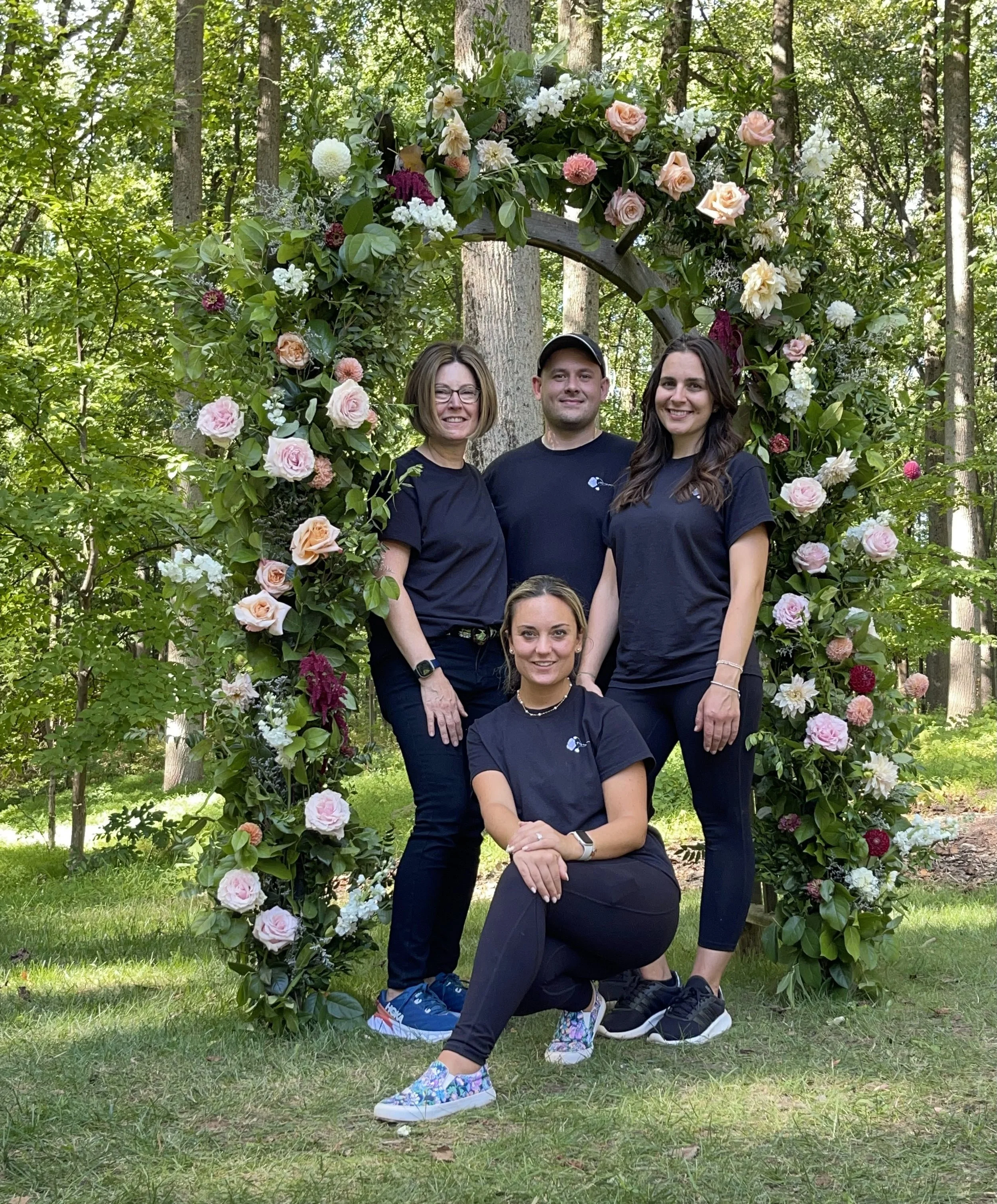 A group of four people posing in front of a floral arch in a wooded area, with three women and one man, all wearing black t-shirts, smiling, and standing on green grass.
