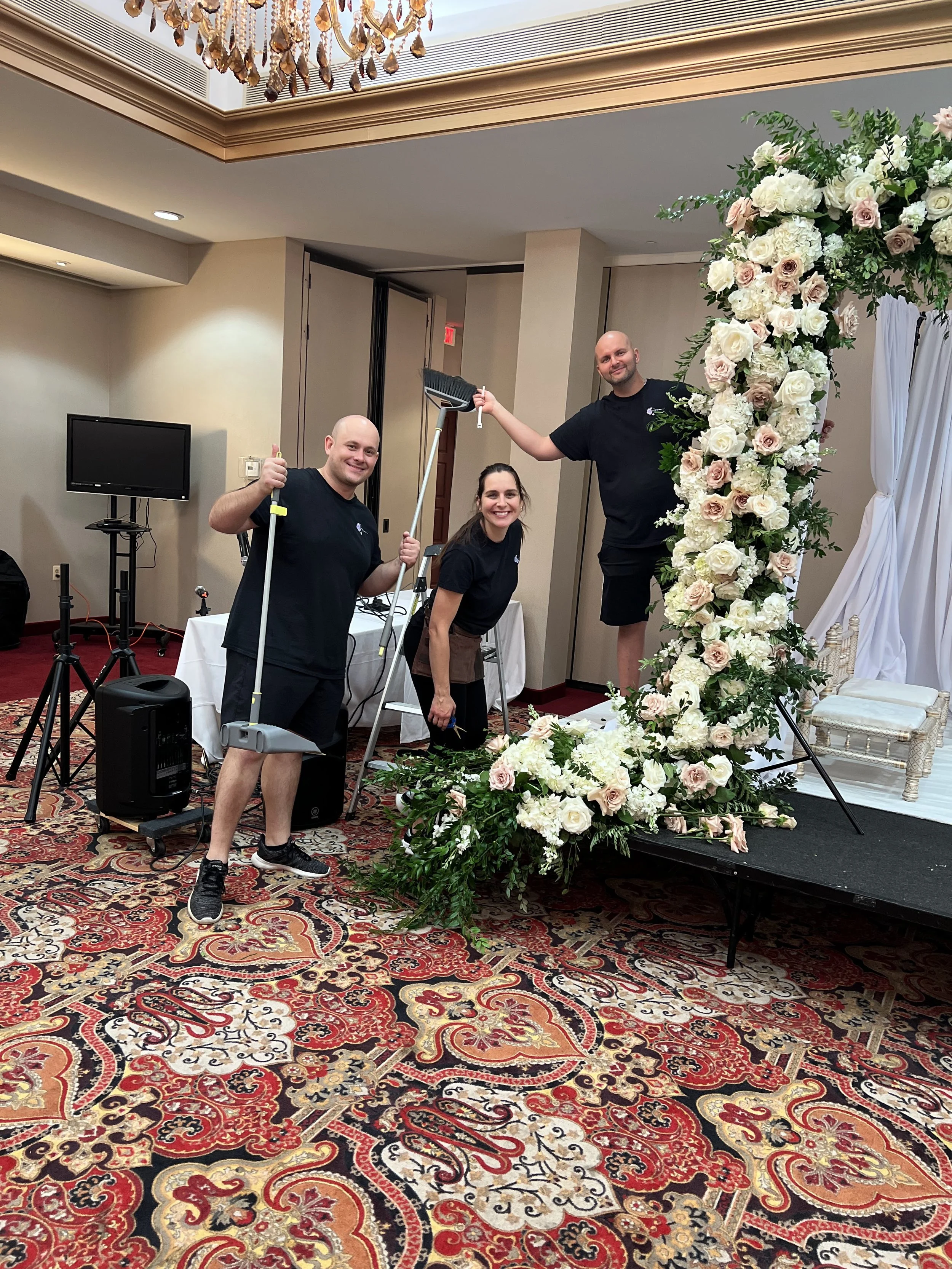 Three people in black shirts standing near a large floral arrangement on a stage at an indoor event. The floral arrangement is made of white and blush roses and greenery. Two people are holding cleaning tools, and all are smiling.