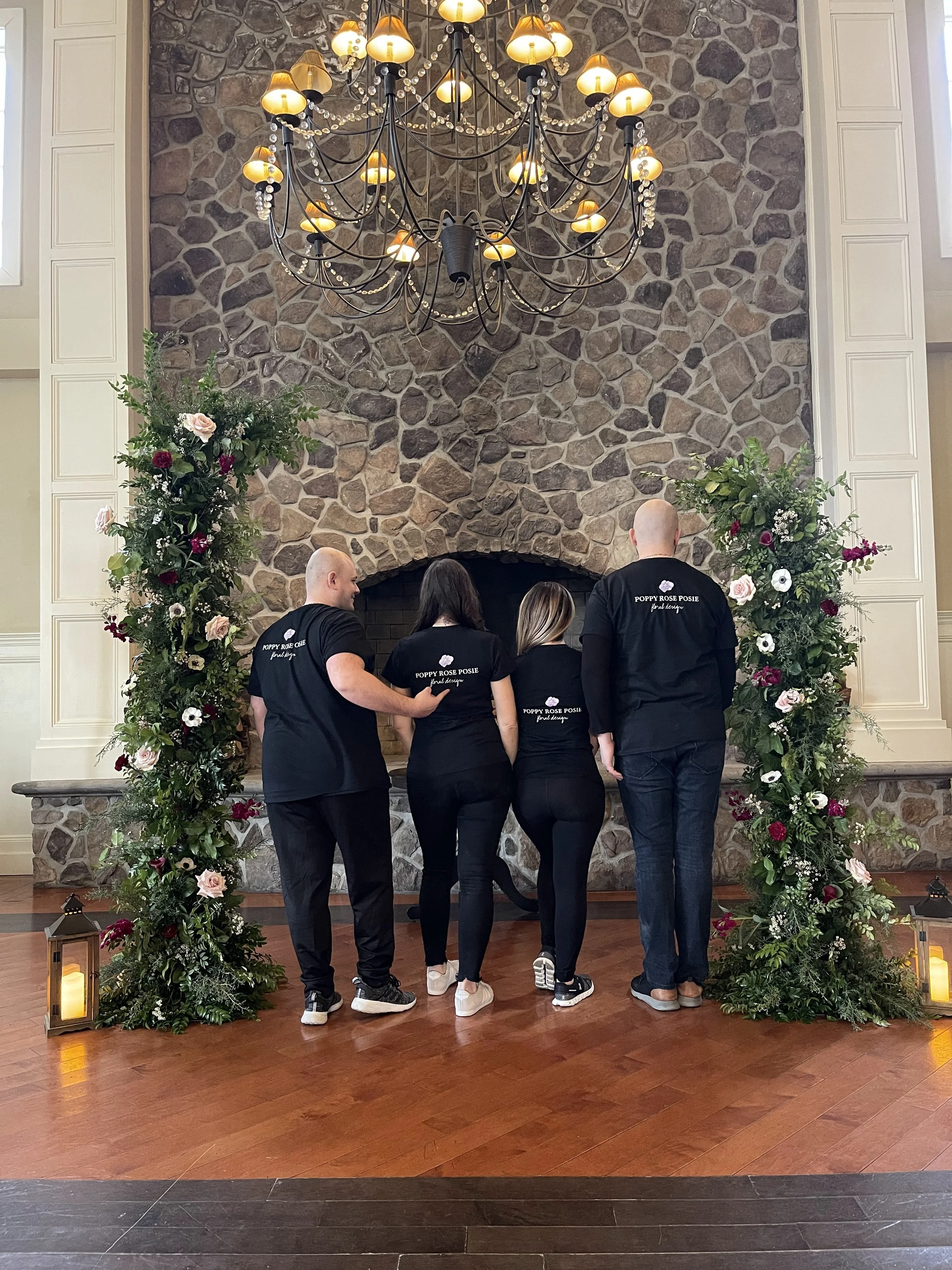 Four people in black shirts standing in front of a stone fireplace, adorned with a large floral arrangement arch and lanterns on the floor, under a big chandelier.