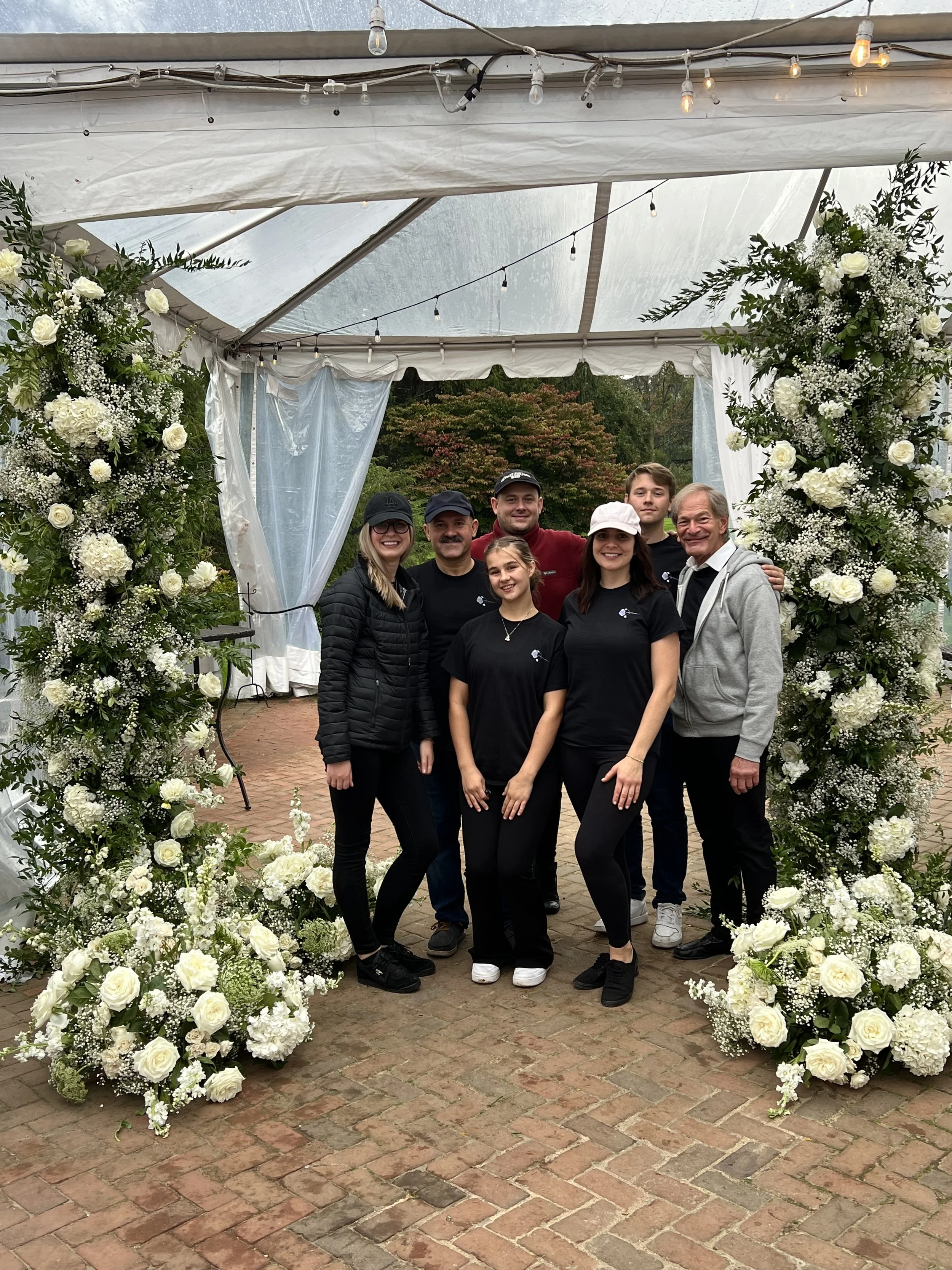 Group of six people standing under an arch decorated with white flowers and greenery, outside with trees in the background.
