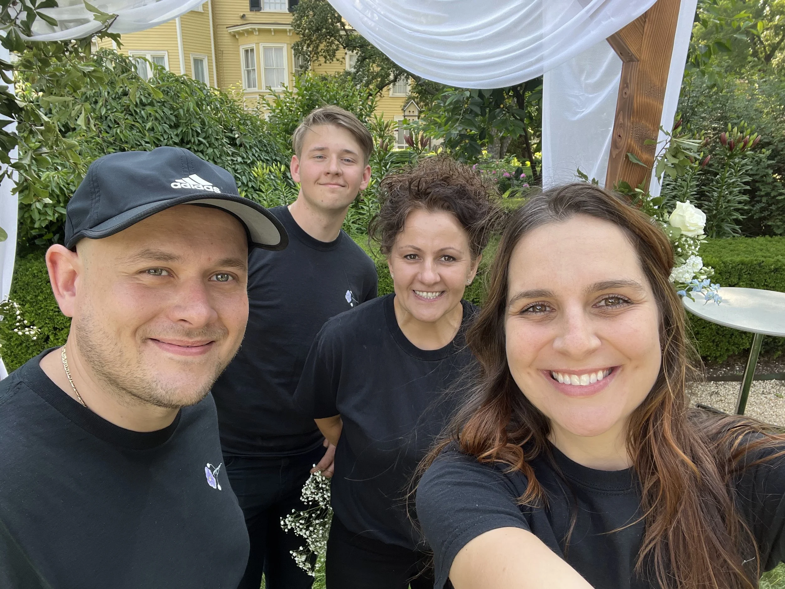 Four smiling people taking a selfie outdoors during daytime. They are standing in front of a garden with greenery and flowers, underneath a wooden and fabric canopy. The group includes two women and two men, all wearing black shirts.