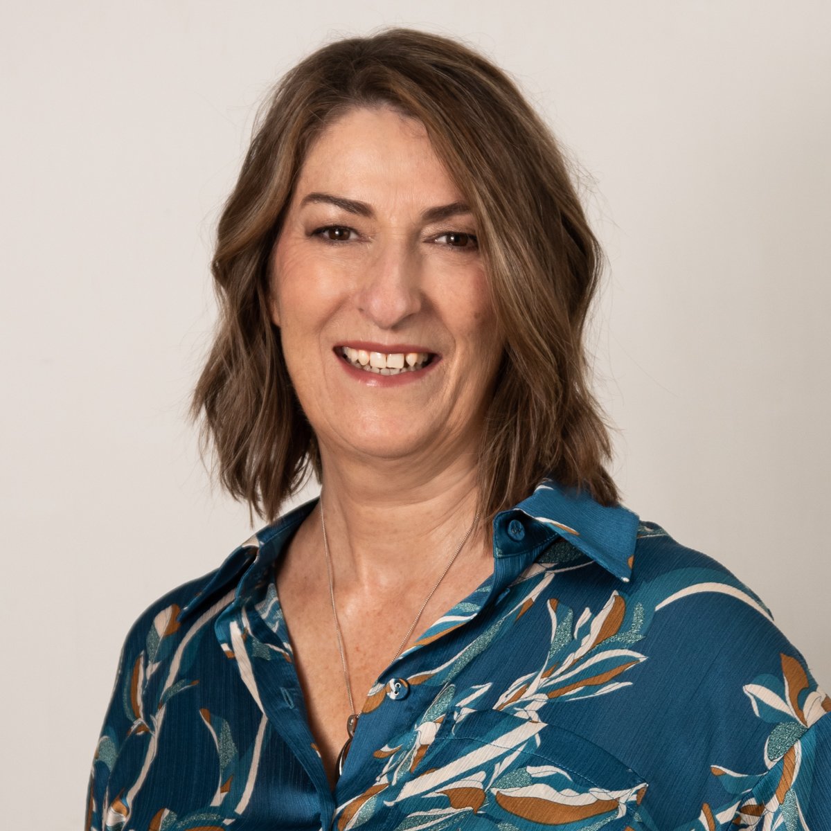 A woman with shoulder-length brown hair smiling, wearing a blue patterned shirt, against a plain light background.