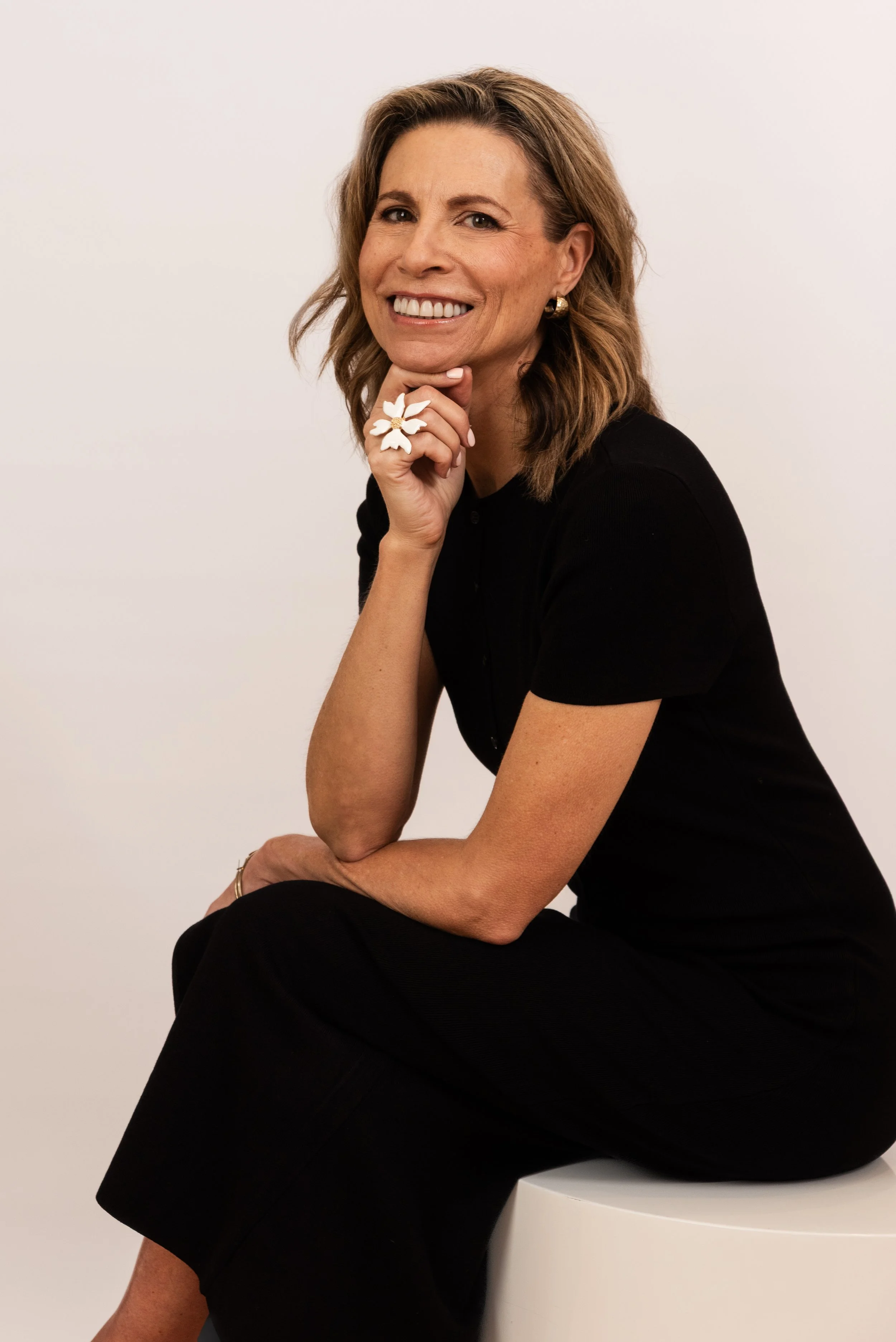 A woman with shoulder-length wavy hair, smiling, wearing a black top, gold hoop earrings, and a large white floral ring, sitting on a white stool against a plain white background.