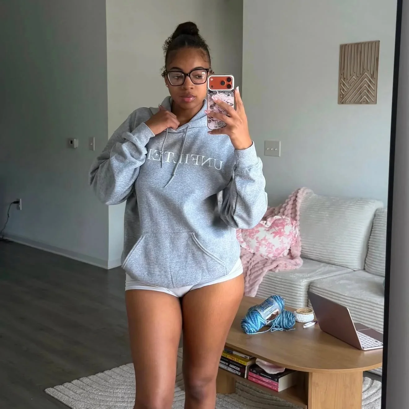 A young woman taking a mirror selfie in a modern living room, wearing glasses, a gray hoodie, and white shorts. There is a sofa with pink and white pillows, a small wooden table with yarn and a laptop, and wall art in the background.