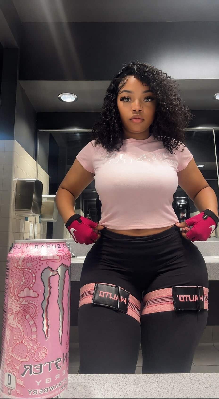 A young woman posing in a gym locker room, wearing black leggings with pink straps, a pink crop top, and pink workout gloves. There is a pink Monster Energy drink can on the counter in front of her.