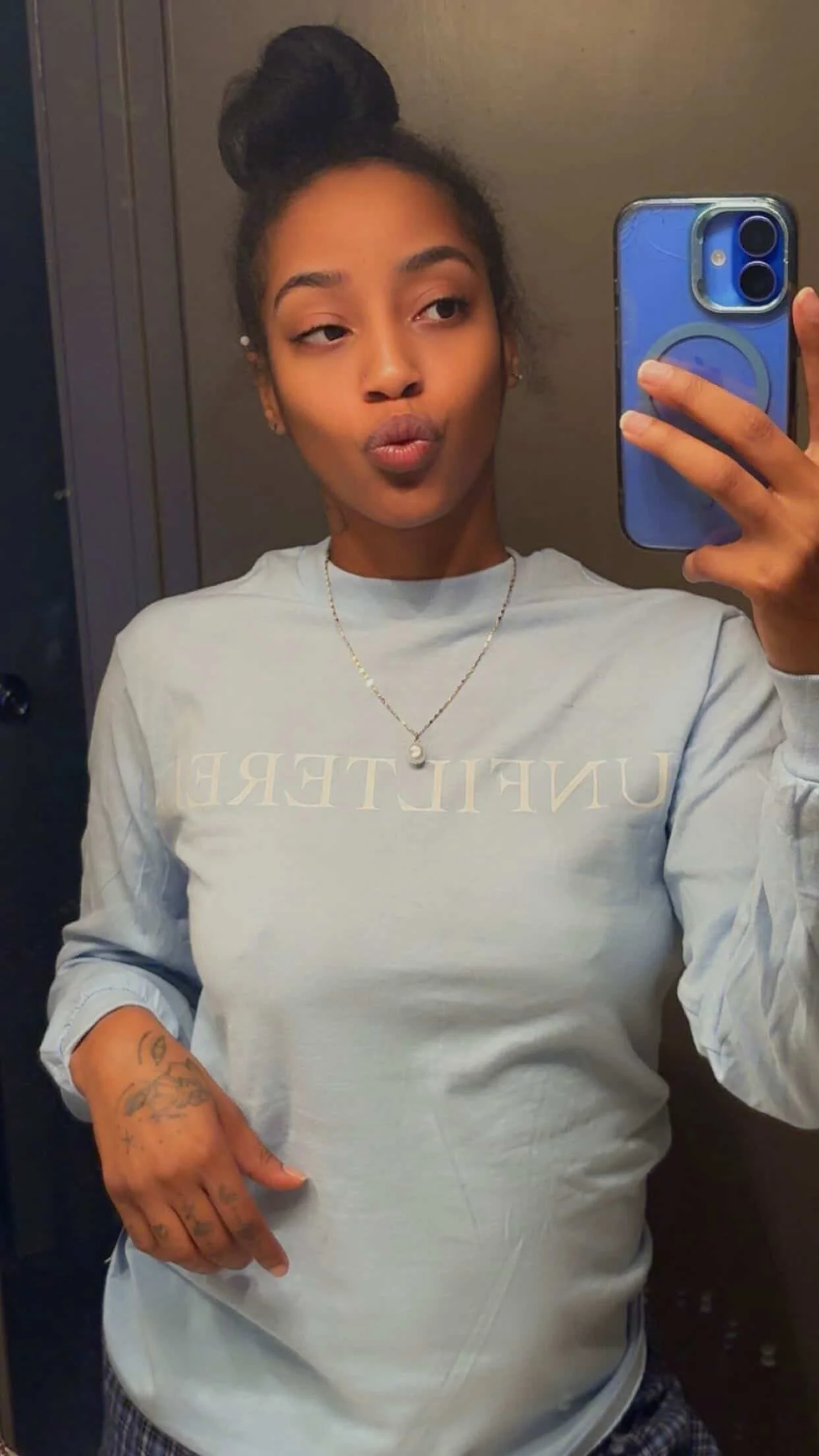 A woman taking a mirror selfie with a smartphone, wearing a light-colored long sleeve shirt, a pearl necklace, and making a puckering face. She has tattoos on her hand and arm, her hair styled in a bun, and is standing in front of a plain wall or door.