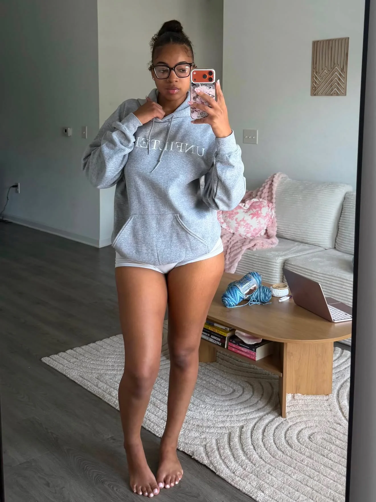 Young woman taking a mirror selfie in a living room, wearing glasses, a gray hoodie, and white shorts, standing barefoot on a wooden floor.