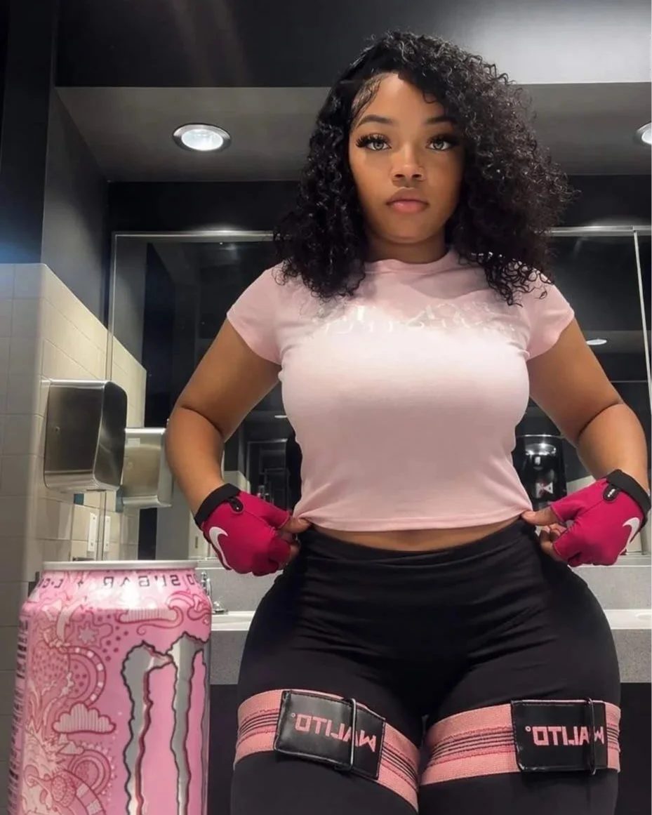 A young woman in workout gear standing in a gym bathroom, holding her waist with both hands. She is wearing a pink t-shirt, black workout shorts, and pink workout gloves. There is a pink Monster energy drink can on the counter beside her.