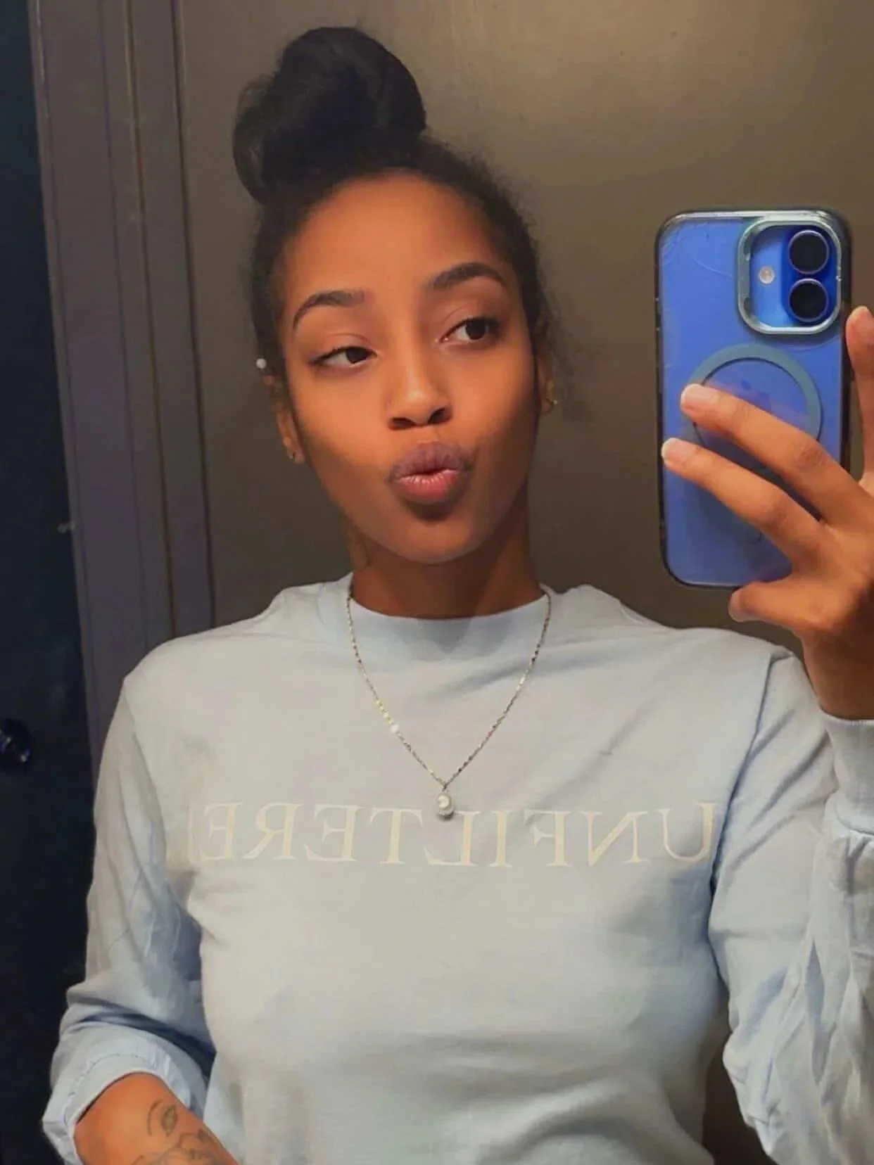 A young woman taking a mirror selfie with a blue smartphone. She has her hair styled in a high bun and wears a light gray sweatshirt with partial text visible. She accessorizes with earrings, a necklace, and has a tattoo on her forearm.