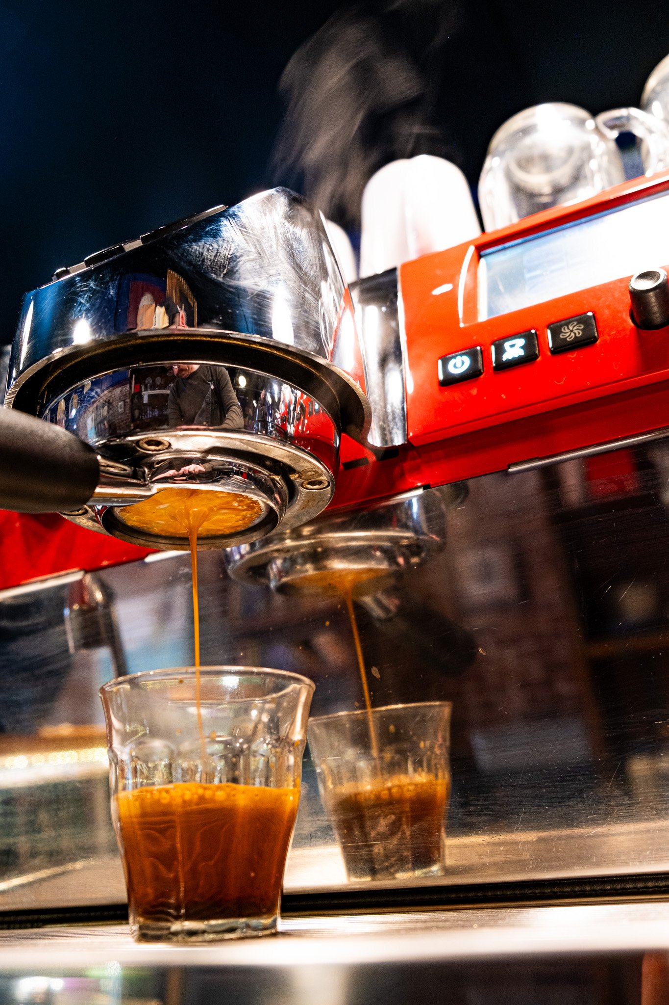 Espresso machine brewing coffee into glass cups, with steam rising and reflections visible on the chrome surface.