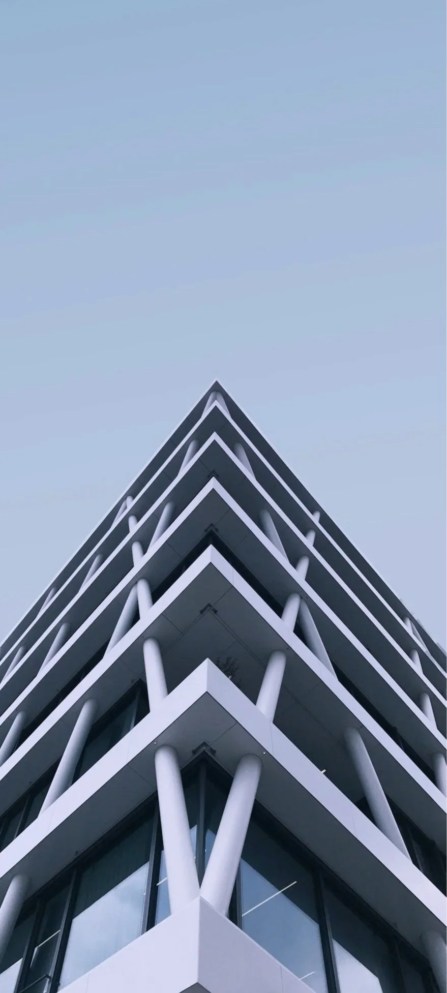 Modern multi-story building with angular design and white structural supports, photographed from below against a light blue sky.