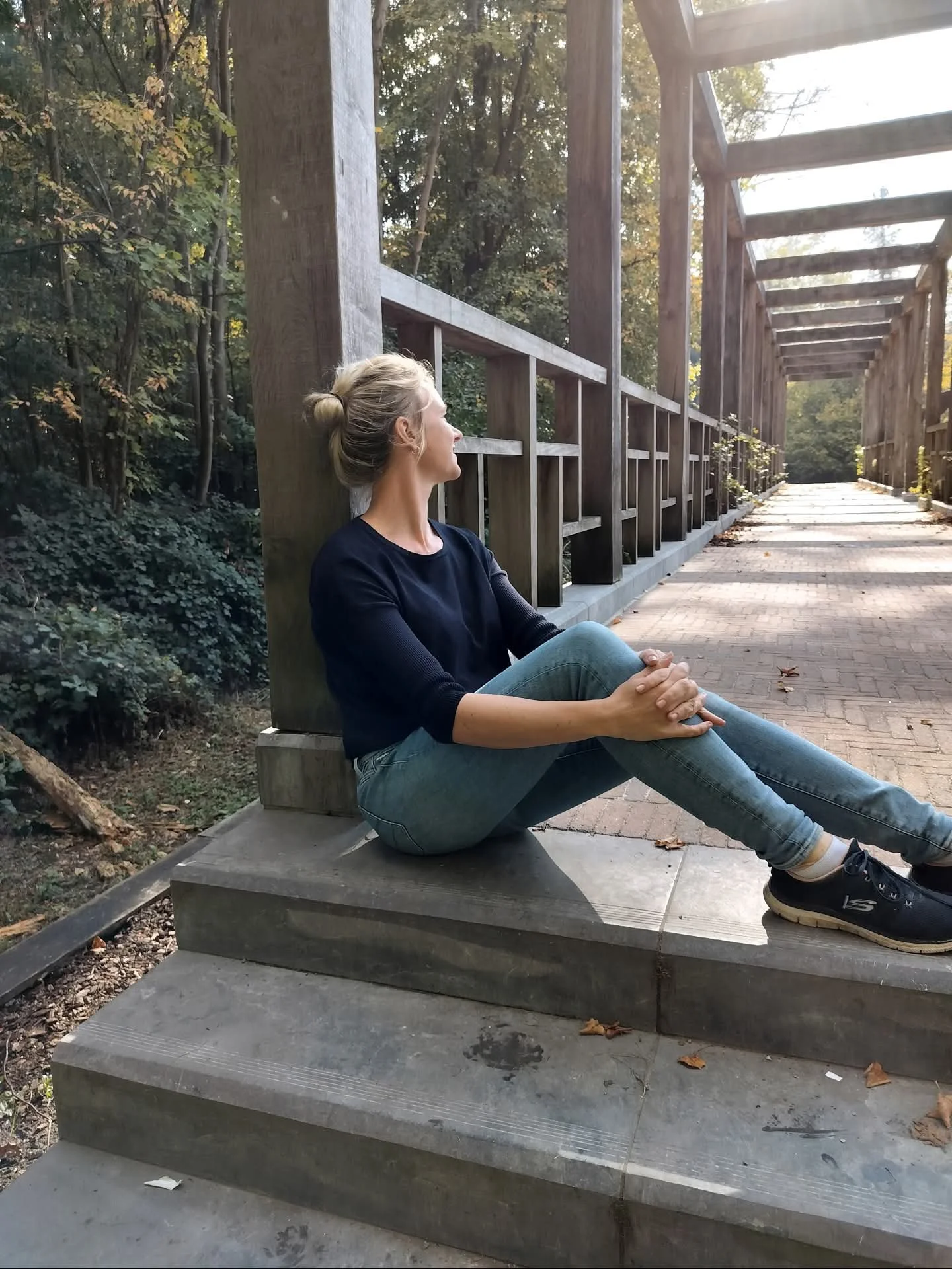 Een vrouw zit op de trappen van een houten brug in een bos, kijkt naar de zon die door de bomen schijnt.