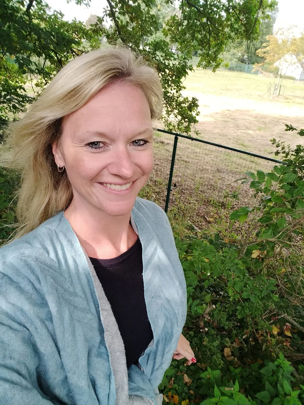 Close-up van een glimlachende vrouw met blonde haar, buiten in een groene tuin met bomen en een hek.