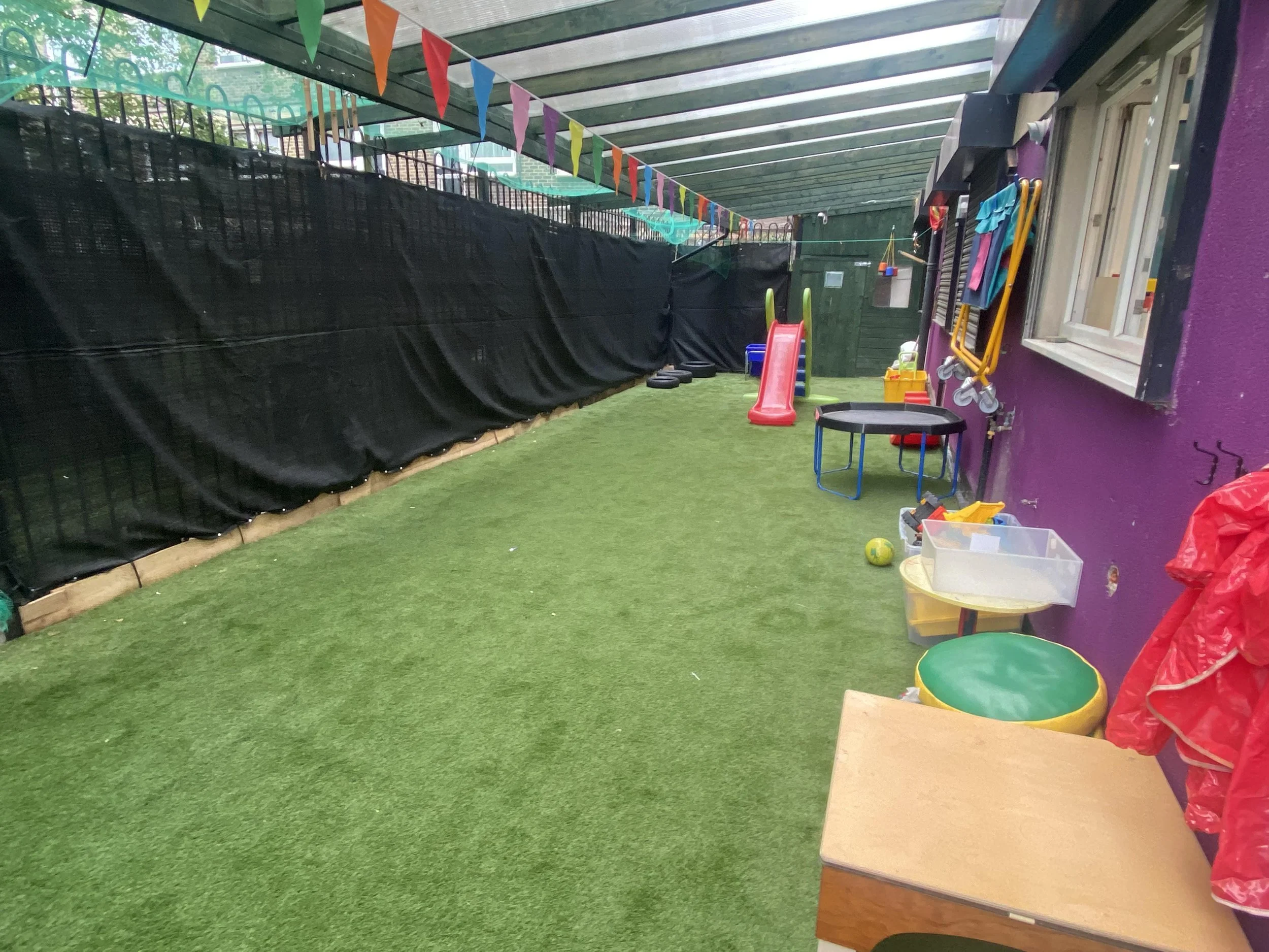 Colorful outdoor children's play area with artificial grass, small slide, trampoline, toys, and playground equipment under a transparent roof.