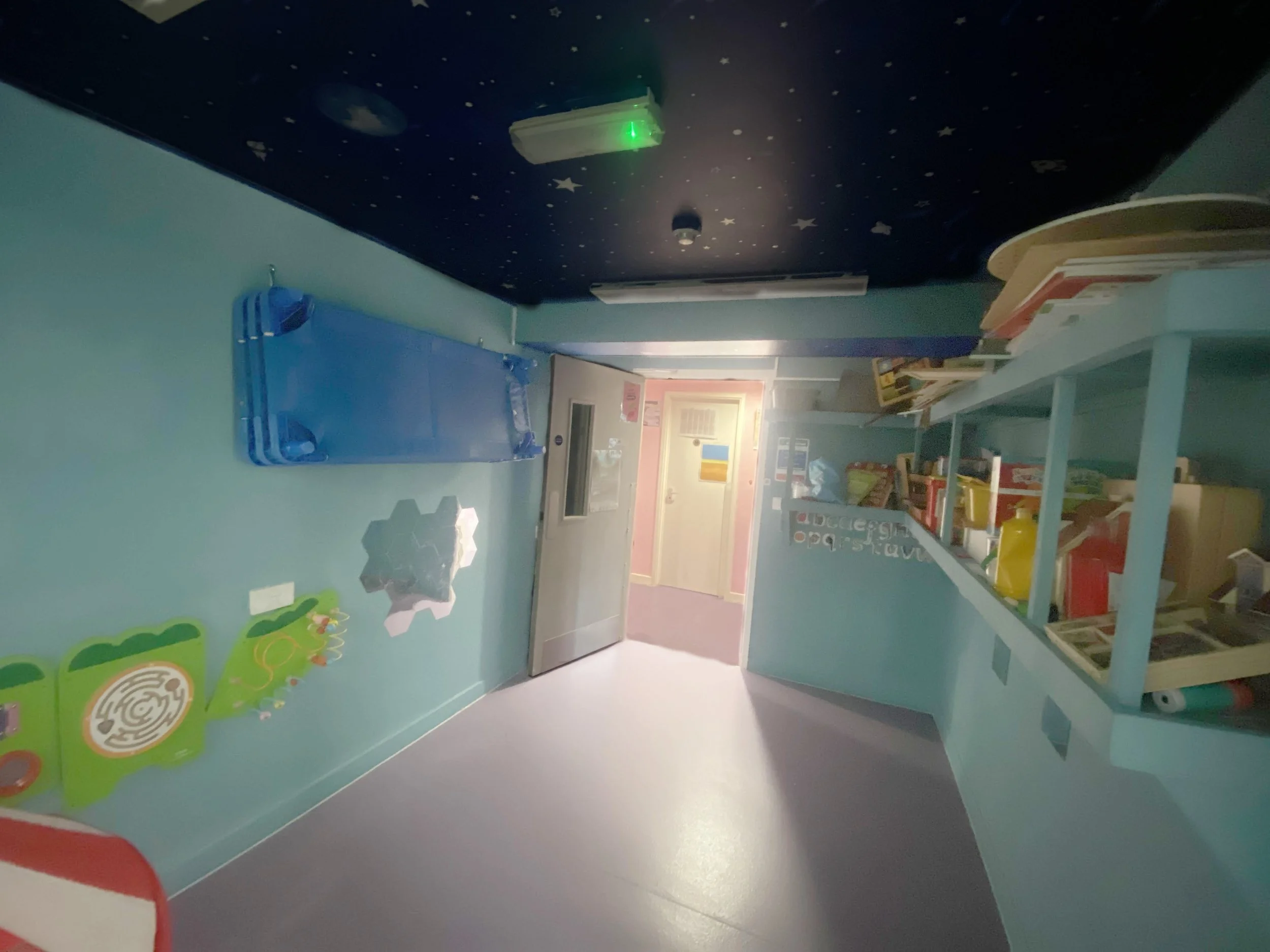 A colorful children's playroom with a teal wall, decorated with wall decals, a blue foldable cot, and shelves filled with toys and books. The ceiling is painted dark with star and moon designs, and a door leads to another room.