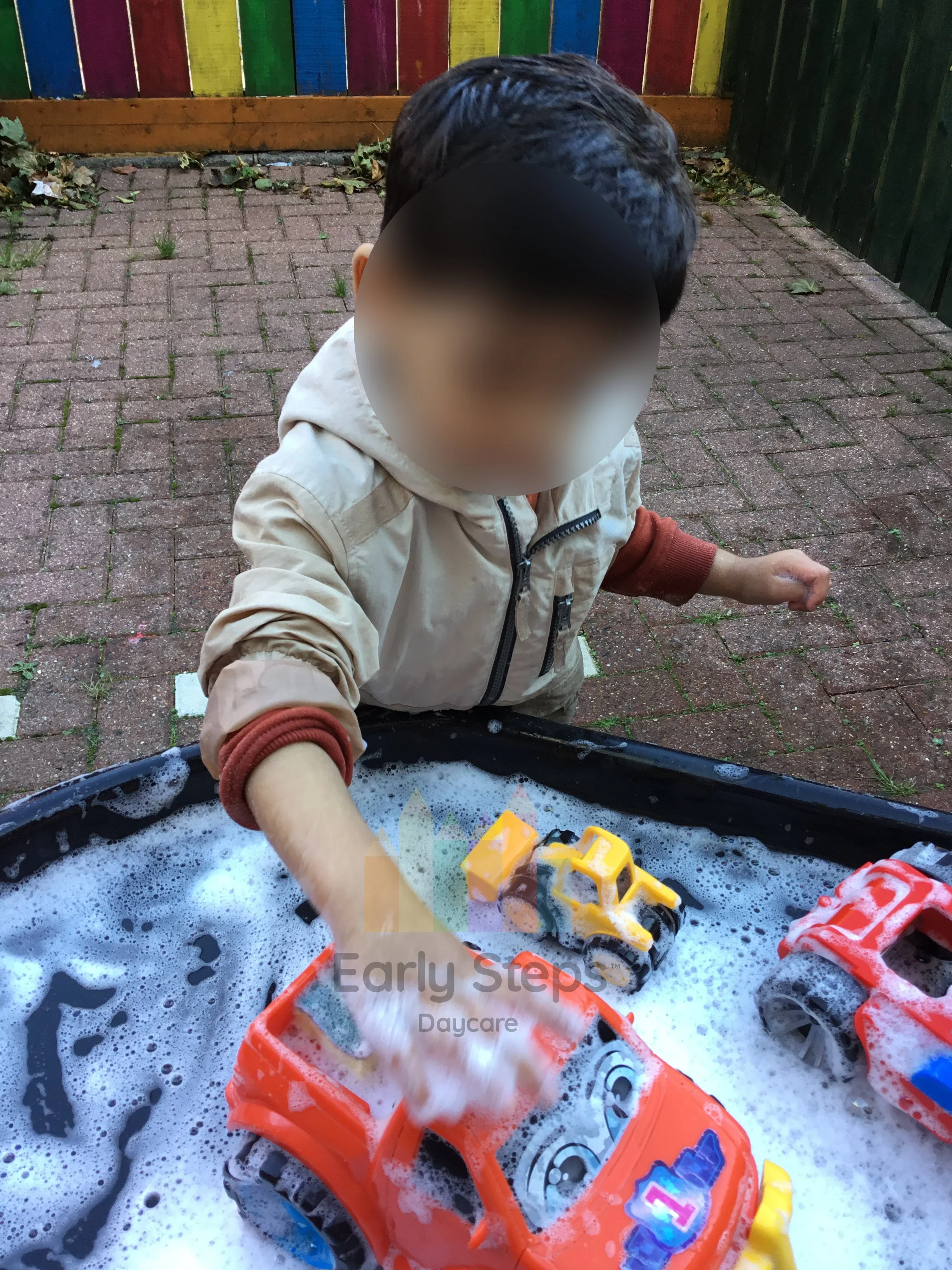 Young child washing toy cars with soapy water and sponges in an outdoor tuff tray, developing fine motor skills and coordination.