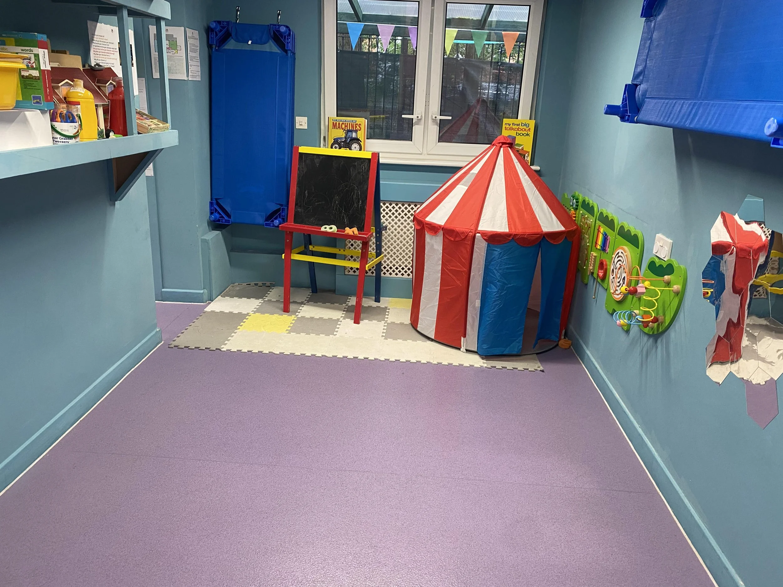 Colorful indoor playroom with a purple floor, a small red and white striped circus tent, a blue chalkboard, a chalkboard with drawing, a green wall-mounted activity panel, and a window with colorful bunting outside.