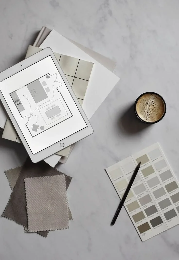 Un tavolo con un tablet, alcuni campioni di tessuto, una palette di colori, una rivista, una penna, una tazza di caffè, alcuni libri e un foglio con campioni di colore.
