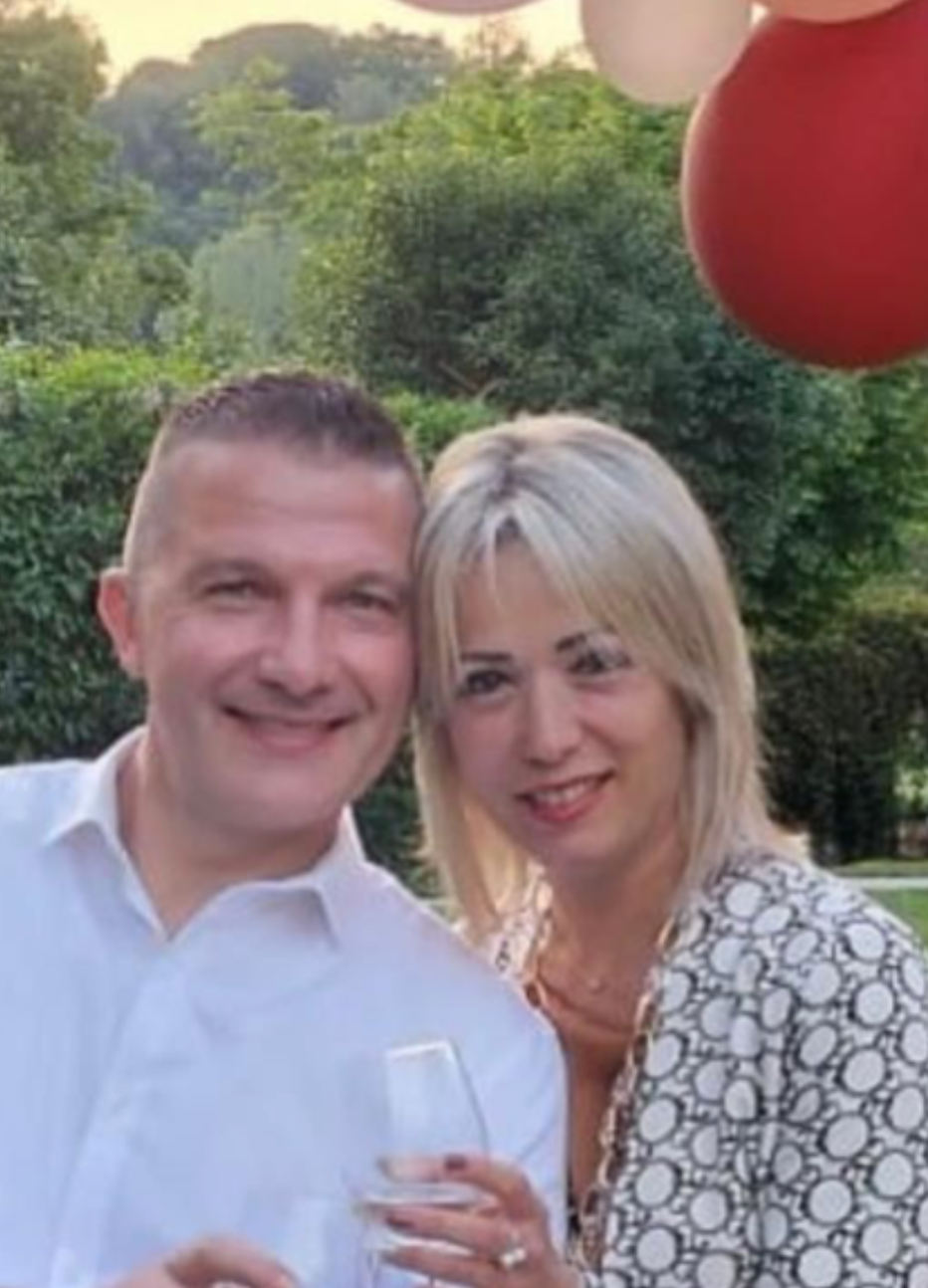 Una coppia sorridente con bicchieri in mano in un giardino, decorato con palloncini rossi e rosa, durante un evento festivo.